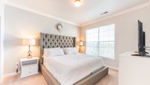 Une chambre à coucher avec un grand lit, deux tables de chevet avec des lampes, un téléviseur posé sur un meuble et une fenêtre avec des stores.
