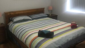 Een net opgemaakt bed met een gestreept sprei, een kussen en een opgerolde handdoek. Daarbij staat een nachtkastje met een lamp naast het bed. Boven het bed hangt een schilderij.
