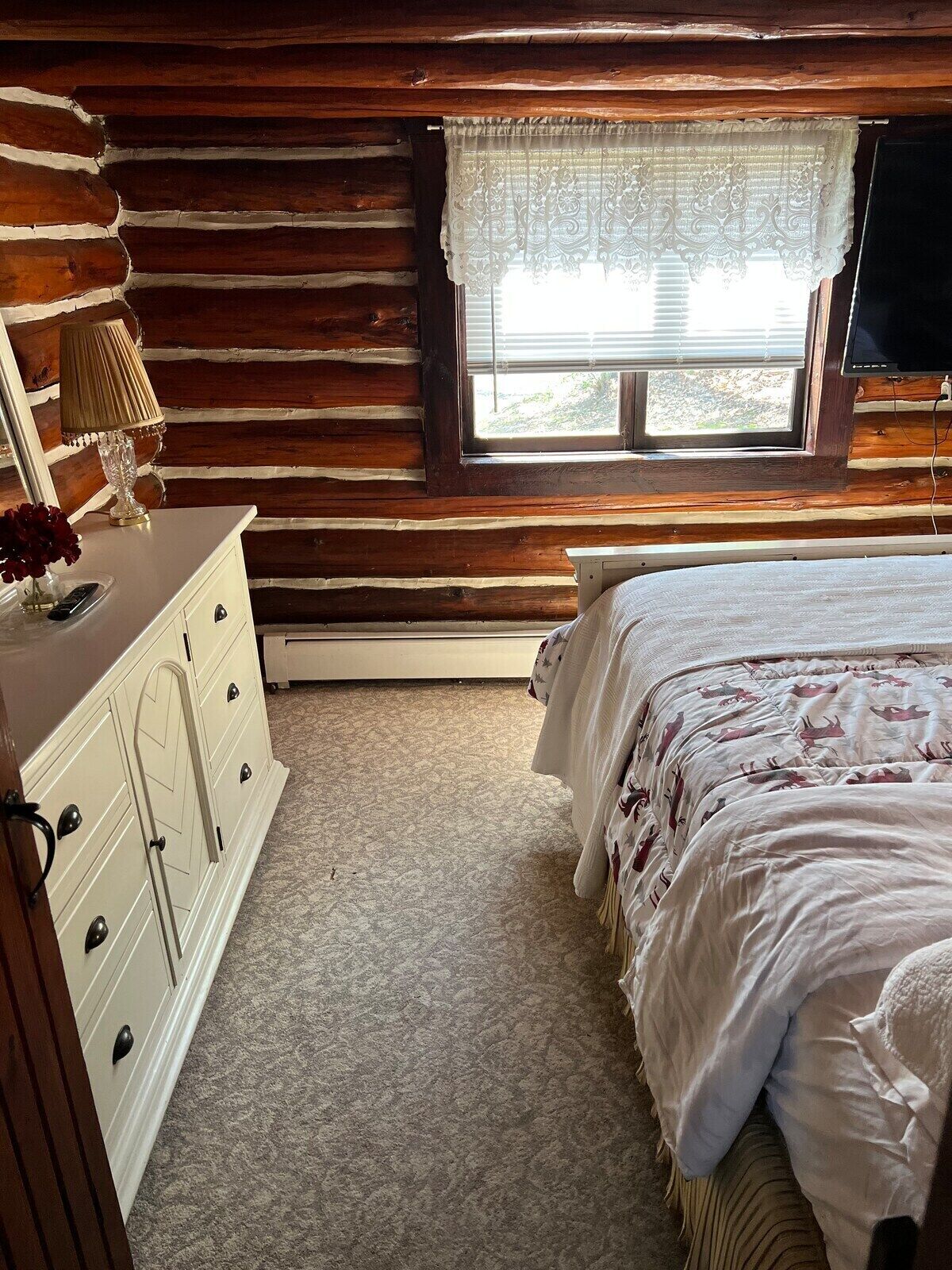 A log cabin bedroom with a bed, dresser, and window with blinds.