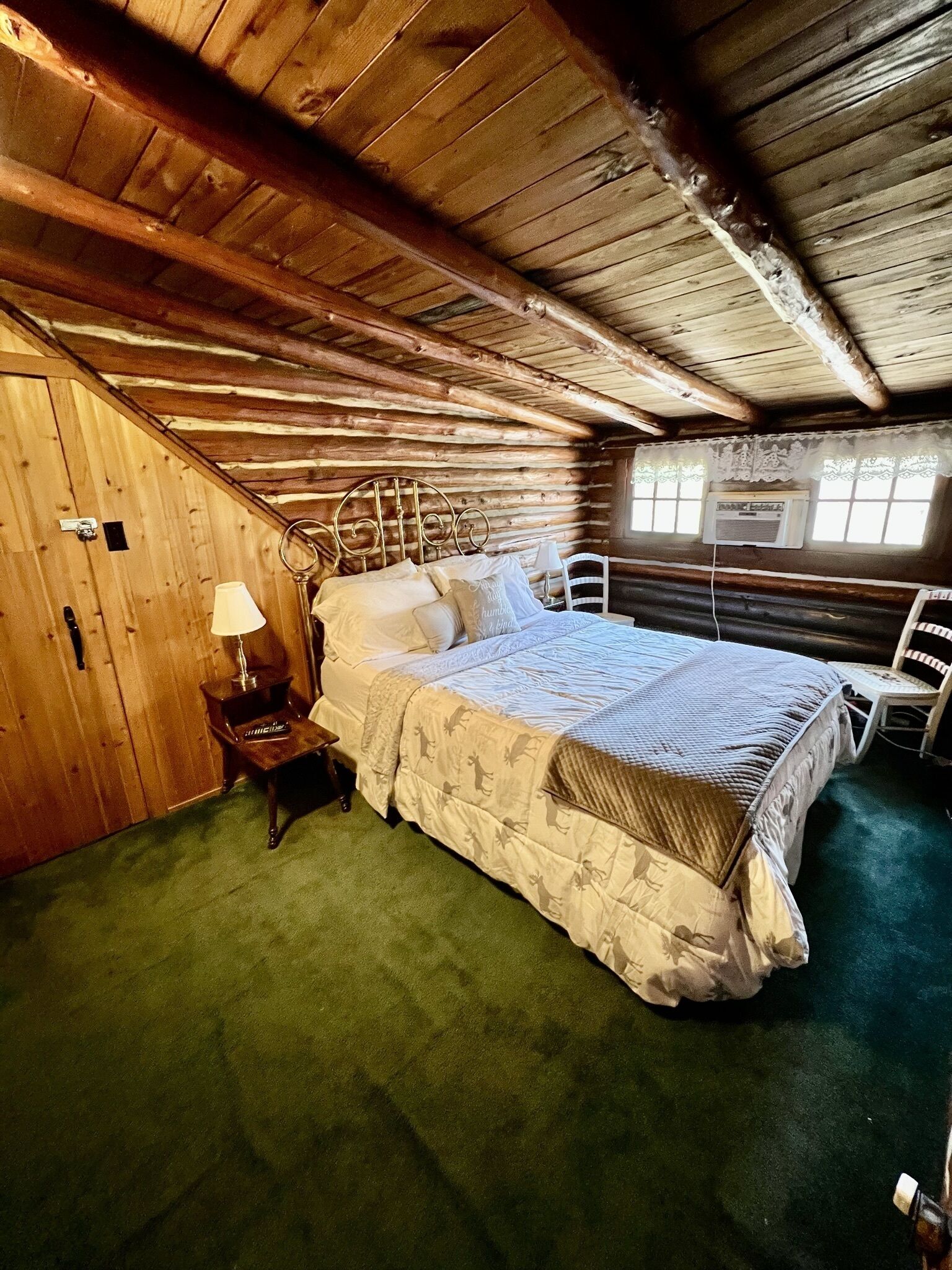 Ein Schlafzimmer in einer Blockhütte mit Bett, Nachttisch, Stuhl und Fenster mit Spitzenvorhängen.