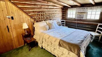 Ein Schlafzimmer in einer Blockhütte mit Bett, Nachttisch, Stuhl und Fenster mit Spitzenvorhängen.