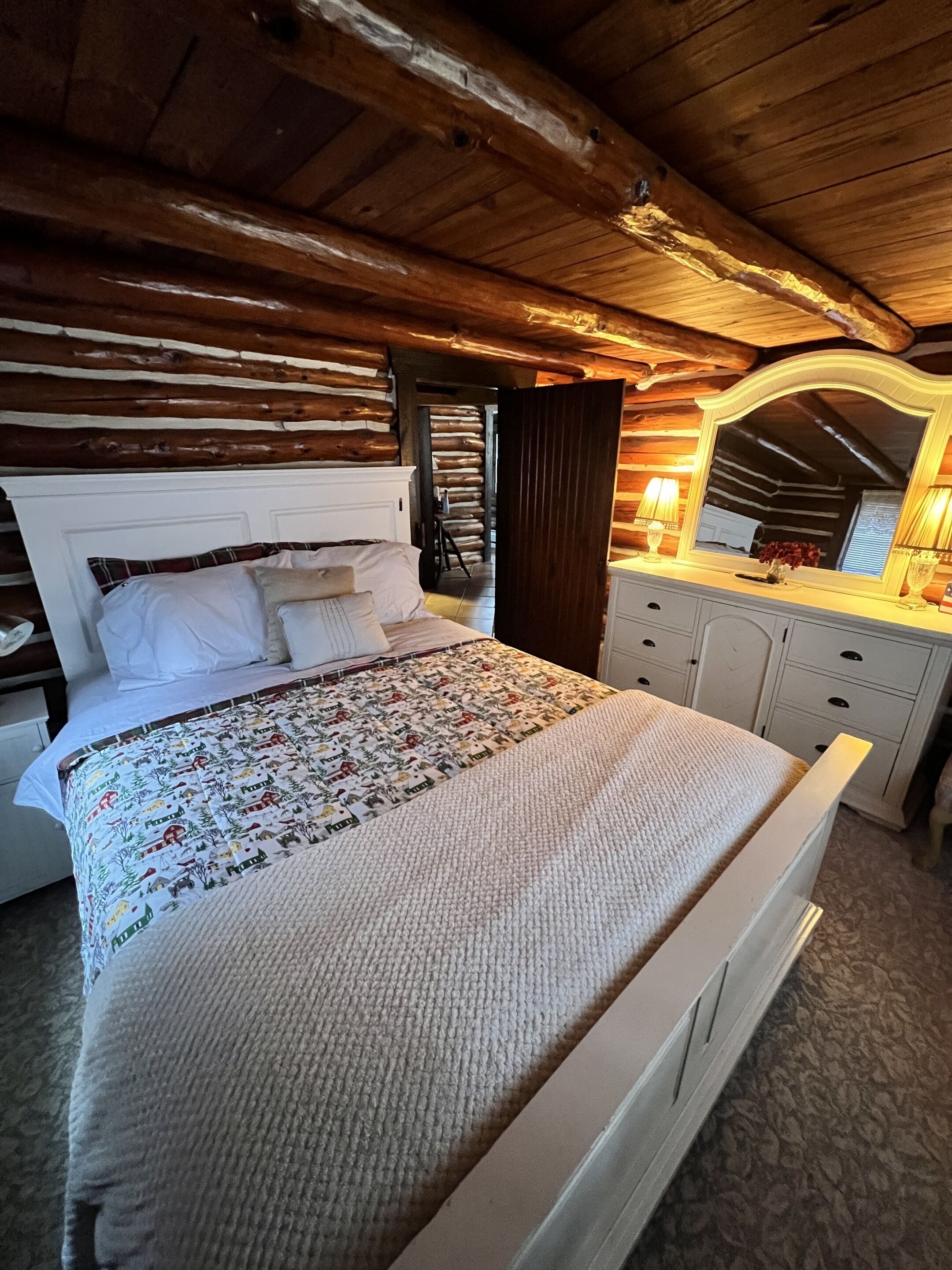 A log cabin-style bedroom with a large bed, wooden ceiling beams, a dresser, and a mirror.
