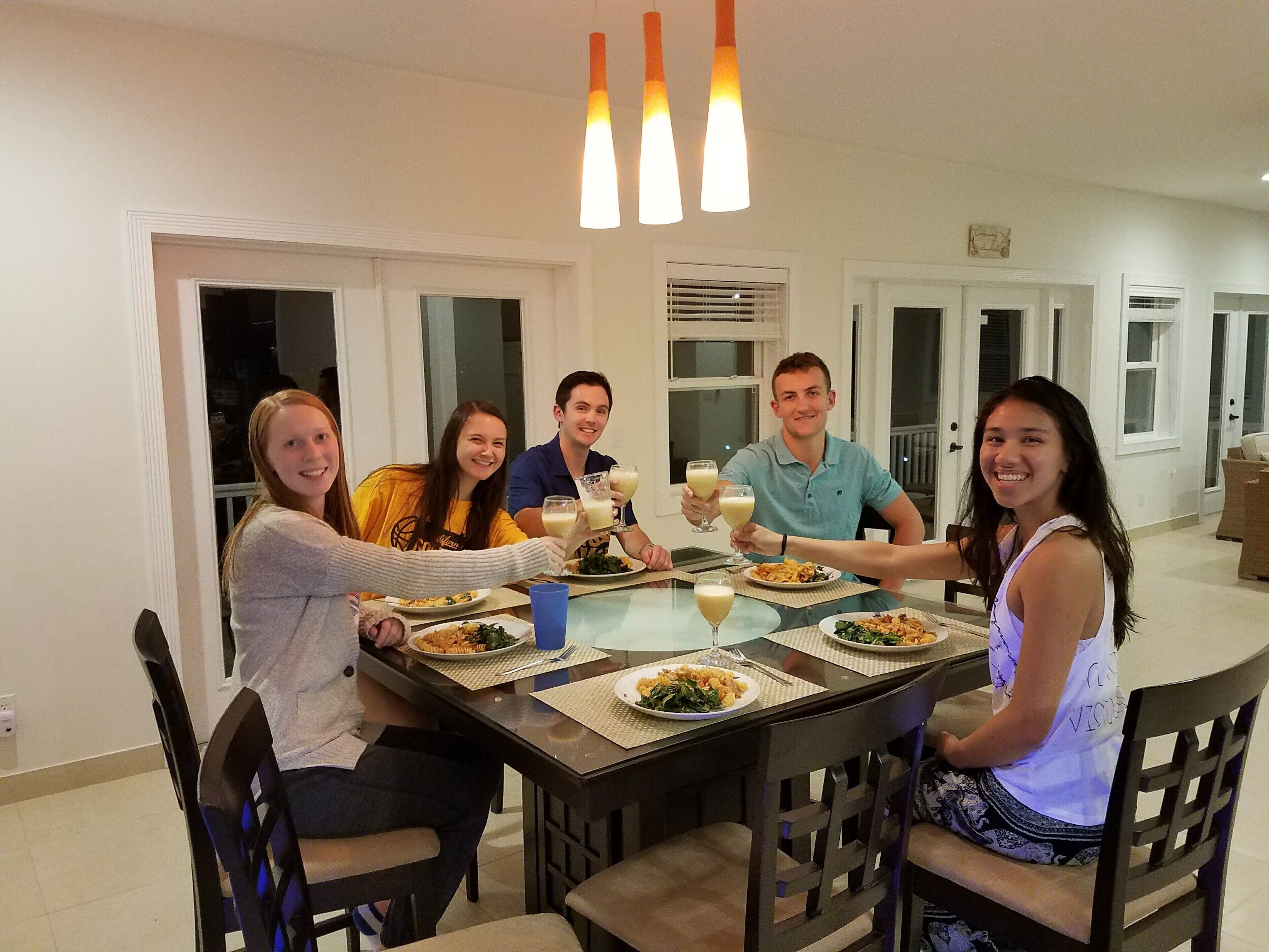 A group of people seated around a dining table in a well-lit room with a modern design, featuring a large window and a ceiling light fixture.