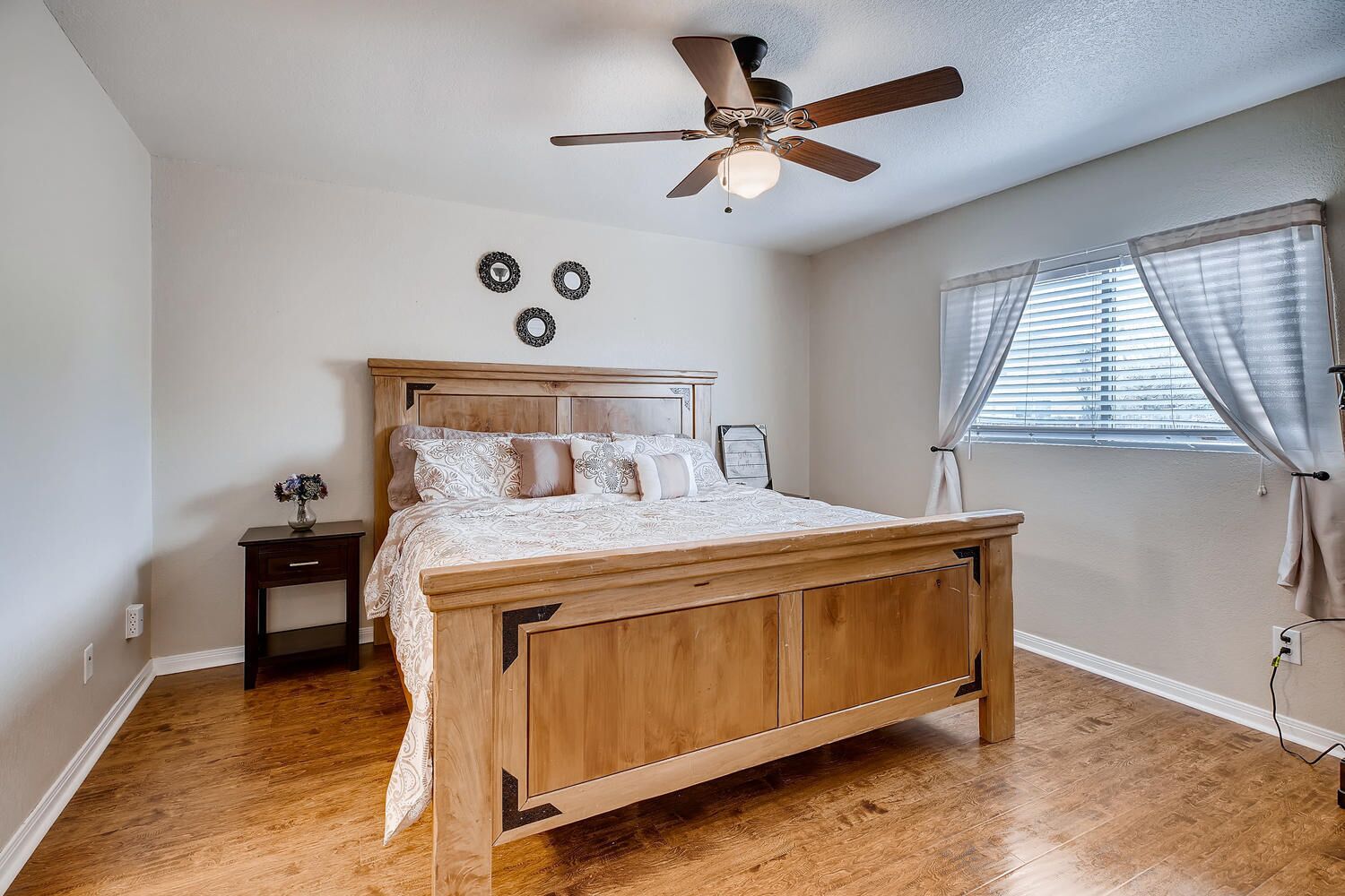 A bedroom with a wooden bed, a ceiling fan, a nightstand, a window with blinds, and a wall-mounted light fixture.