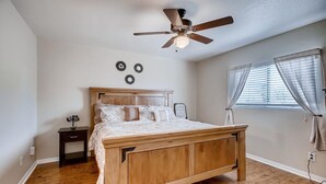 Un dormitorio con cama de madera, ventilador de techo, mesita de noche, ventana con persianas y lámpara fijada a la pared.