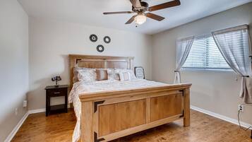 A bedroom with a wooden bed, a ceiling fan, a nightstand, a window with blinds, and a wall-mounted light fixture.