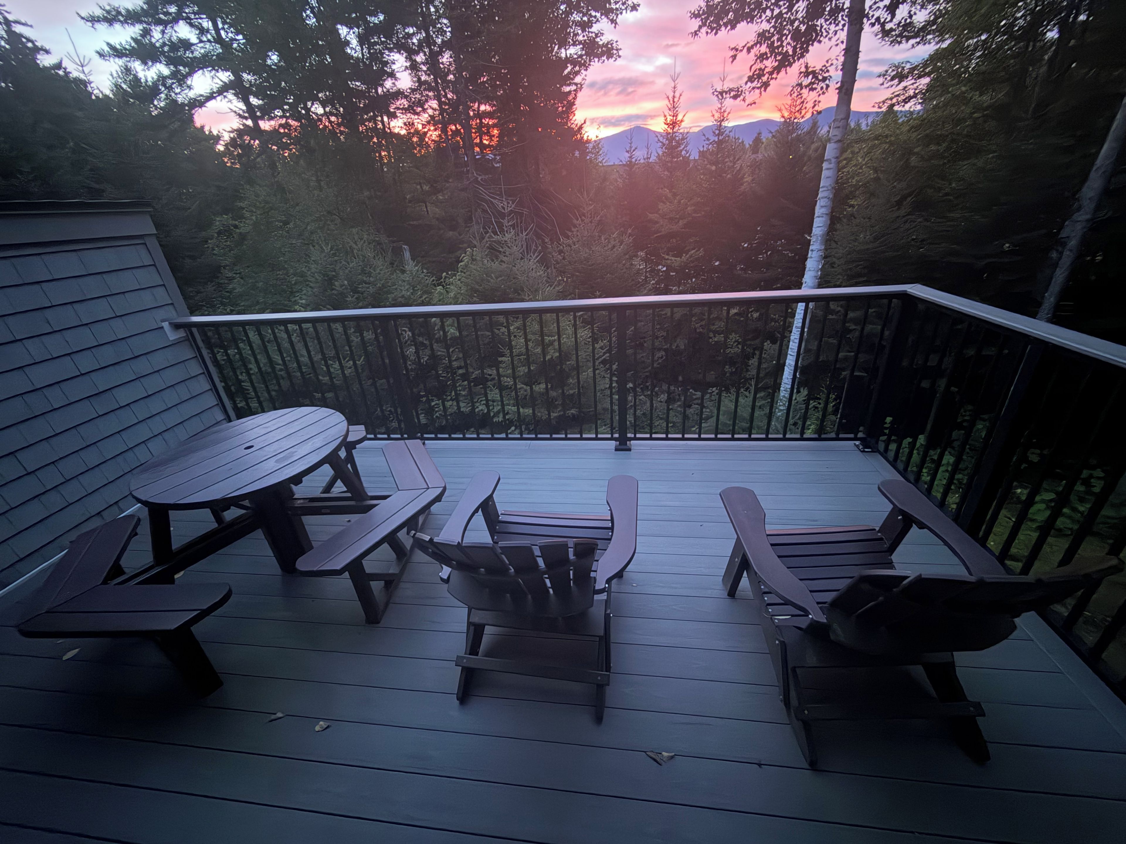 Una terraza de madera con mesas de picnic y bancos, rodeada de árboles y vistas al atardecer.