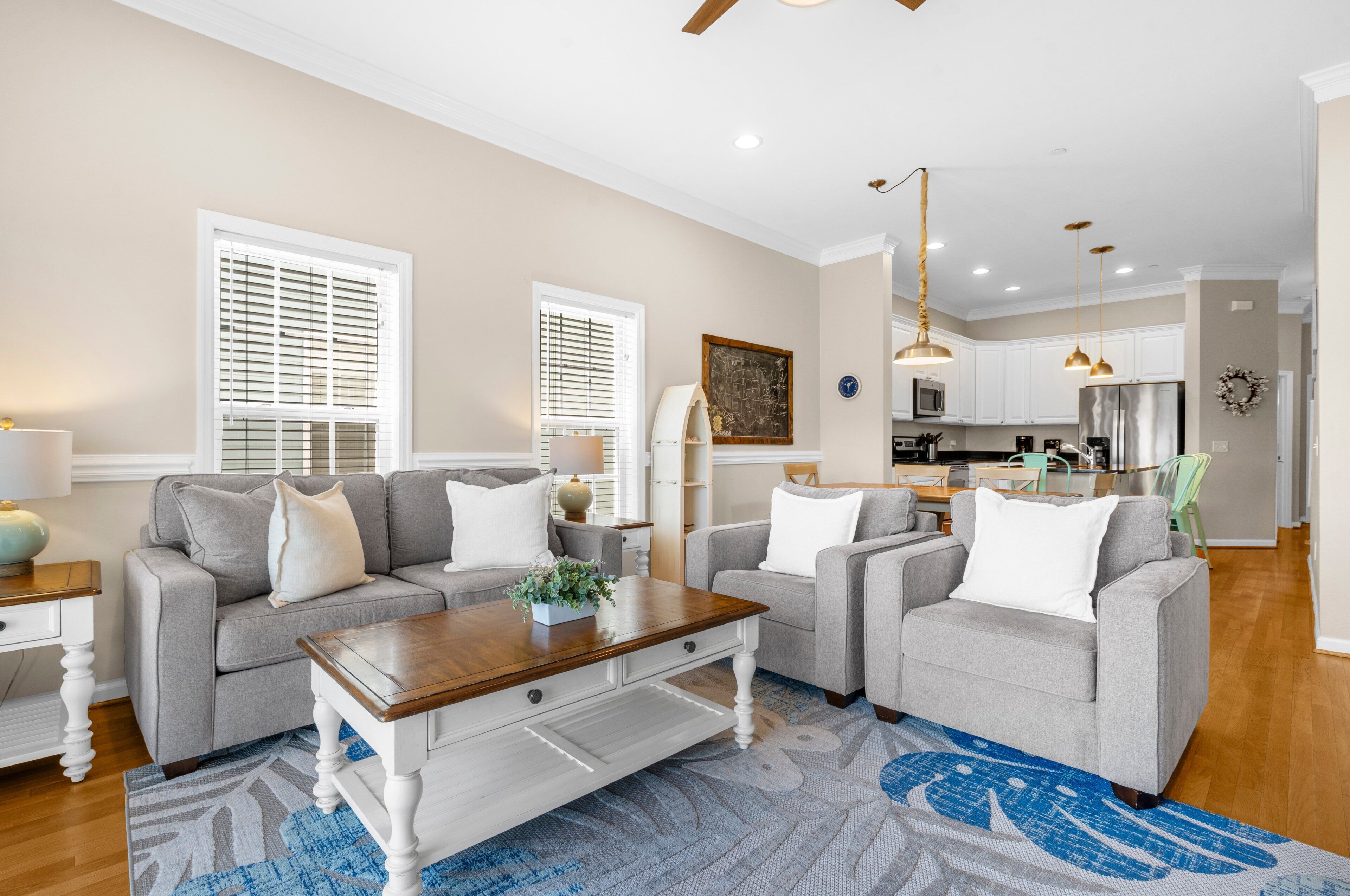 A living room with a sofa set, a coffee table, and a rug. There is a kitchen in the background with cabinets and a refrigerator.