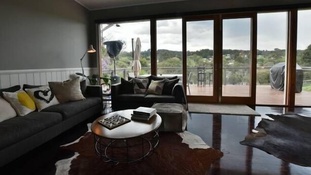 A modern living room with a sofa, a coffee table, and a view of a balcony with outdoor furniture. - Waterside Cottage - Perfect Lakeside Location (Daylesford)