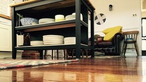 A room with a wooden floor, a dark-colored bookshelf with plates, a yellow cushion, and a small chair. - Waterside Cottage - Perfect Lakeside Location (Daylesford)