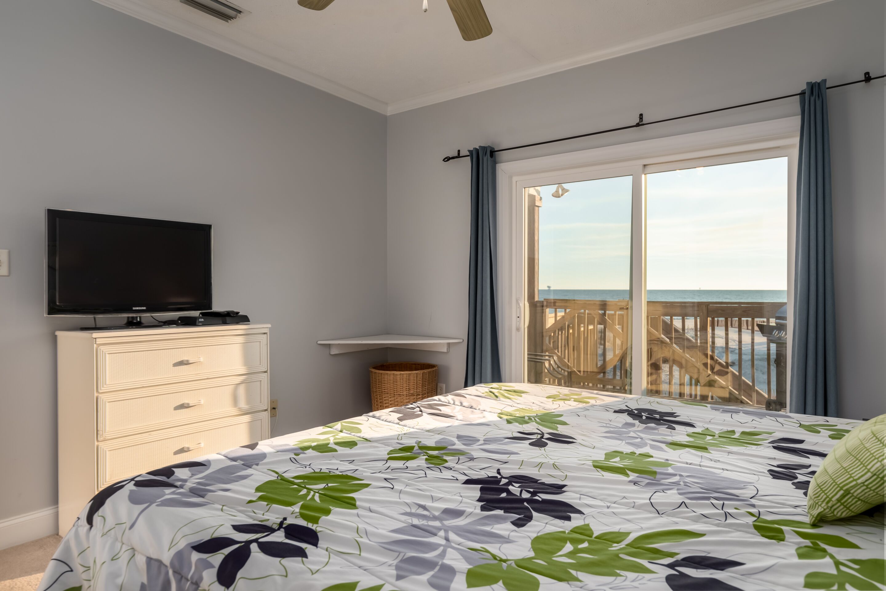 A bedroom with a bed, a TV on a dresser, a balcony with a view of the sea, and a wooden railing.