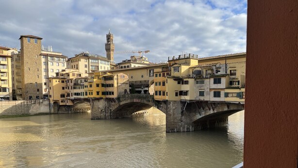 Un pont historique, avec des arches en pierre enjambant une rivière, est entouré d’immeubles à plusieurs étages dotés de balcons et d’une tour.