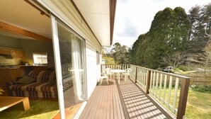 A wooden deck with a dining set and a sliding glass door leading to a living area with a sofa and a table. - Grevillea Cottage - Family and Group Getaways (Katoomba)