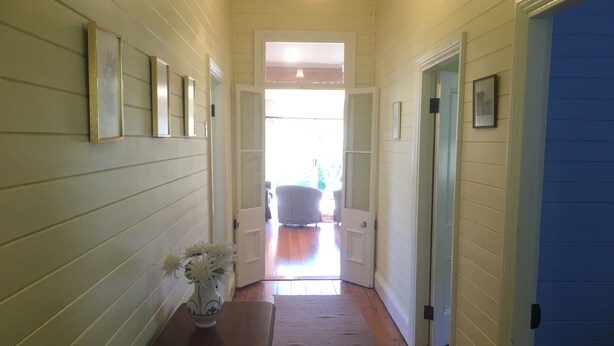 A hallway with wooden paneling, a small table with a vase, and a door leading to a bright room with a chair. - Grevillea Cottage - Family and Group Getaways (Katoomba)