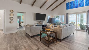 A spacious living room with a large sofa, a coffee table, and a flat-screen TV. There is a sliding glass door leading to a balcony with a view of the ocean. - The nicest beach house in Great Harbour,   Guaranteed!   Enjoy🏝 (Great Harbour Cay, Bahamas (GHC))