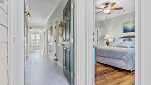 A hallway leading to a bedroom with a ceiling fan, a bed with a 'SUMMER' pillow, and a view of the ocean through the window. - CottageVacations4u "MEANT 2B" -pool club- ocean view-200 FT to beach. DOG FRIENDLY. (Ocean Isle Beach)