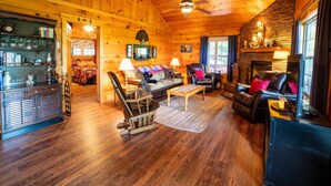 A cozy log cabin interior with a fireplace, leather sofas, a wooden coffee table, and a bookshelf. - Haven in the Hills! Spectacular Views, River Access, & 5-Star Amenities! (Murphy)