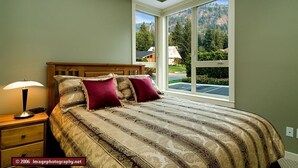 A bedroom with a large window, a wooden headboard, a patterned bedspread, and a bedside lamp.