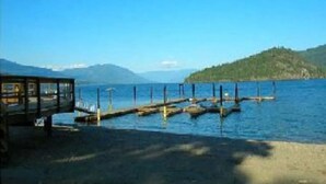 A lakeside dock with a wooden structure on the left, a clear blue sky, and a distant mountain range.