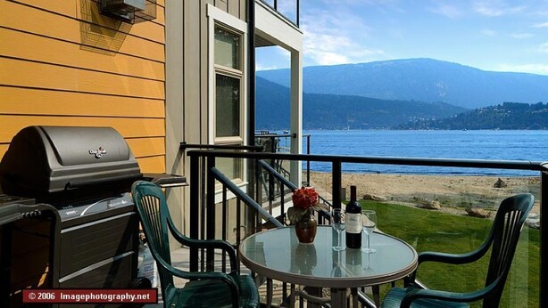 A balcony with a grill, chairs, and a table with a bottle and glass, overlooking a beach and mountains.
