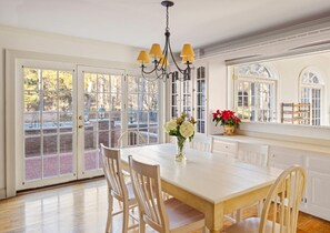 Un coin repas avec une table et des chaises blanches, un lustre et une vue dégagée grâce à de grandes portes vitrées.
