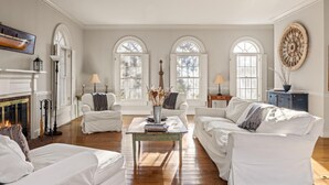 Un salon spacieux, meublé de blanc, doté de grandes fenêtres et d’une cheminée.