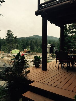 Eine Holzterrasse mit Tisch und Stühlen, umgeben von Blumentöpfen und Blick auf einen bewaldeten Hügel.