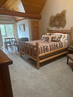 A wooden-framed bed with patterned bedding, a rustic wall decor, and a view of the outdoors through a large window.