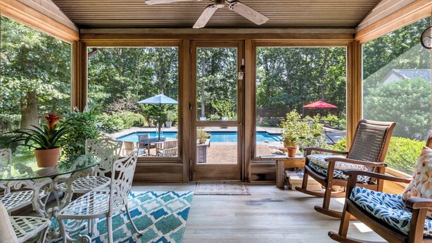 A covered patio with a ceiling fan, a glass table, white metal chairs, a rocking chair, and a view of a swimming pool and garden.