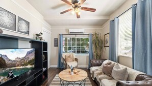 A living room with a television, a ceiling fan, a sofa, a chair, and a window with blinds. - 1890s Gem | Prime Midtown Hub | Walk to Everything (Sacramento)