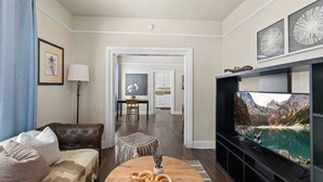A living room with a leather sofa, a round coffee table with a plant, a TV with a mountainous landscape, and a dining area in the background. - 1890s Gem | Prime Midtown Hub | Walk to Everything (Sacramento)