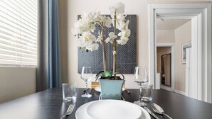 A dining table set with white plates, cutlery, and two wine glasses. A vase with white flowers is on the table, and a geometric wall art is visible in the background. - 1890s Gem | Prime Midtown Hub | Walk to Everything (Sacramento)