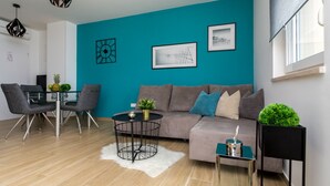 A modern living room with a teal accent wall, a grey sofa, a glass dining table with chairs, and a coffee table with a plant.