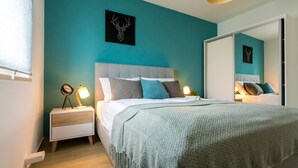 A modern bedroom with a teal wall, a large bed with a grey headboard, and a nightstand with a lamp.