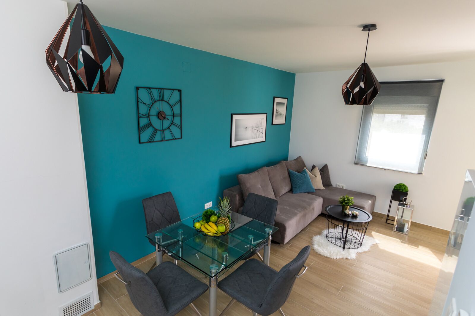Ein modernes Wohnzimmer mit einer türkisfarbenen Akzentwand, einem Glastisch mit Stühlen, einem Sofa und zwei Pendelleuchten.