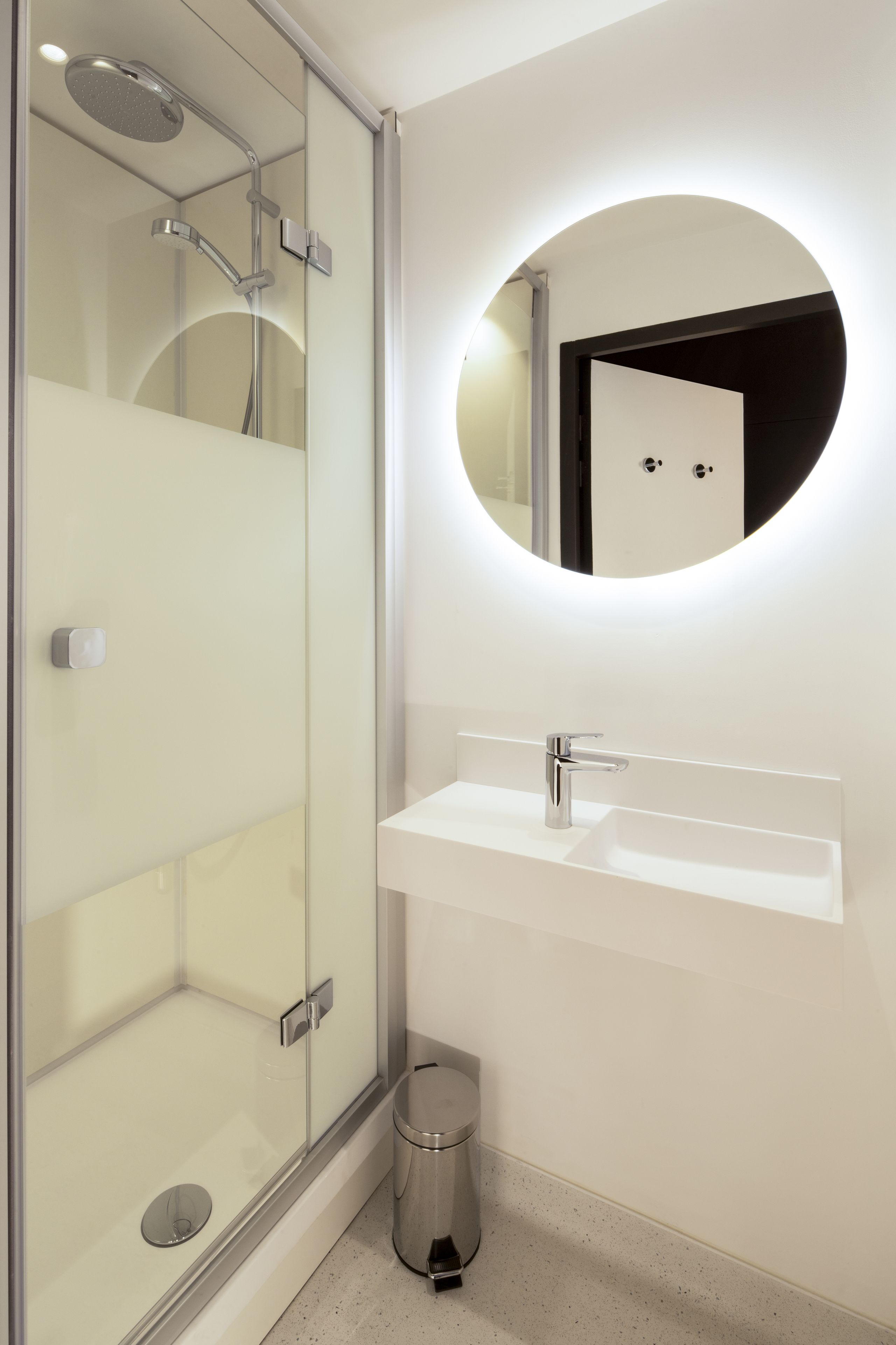 A modern bathroom with a glass-enclosed shower, a white sink, and a round mirror with integrated lighting.