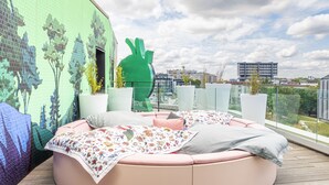 A round, pink cushioned seating area with floral bedding, a green abstract sculpture, and a mural of trees on a balcony with a city view. - JO&JOE Paris - Gentilly Hotel - Hostel (Gentilly)
