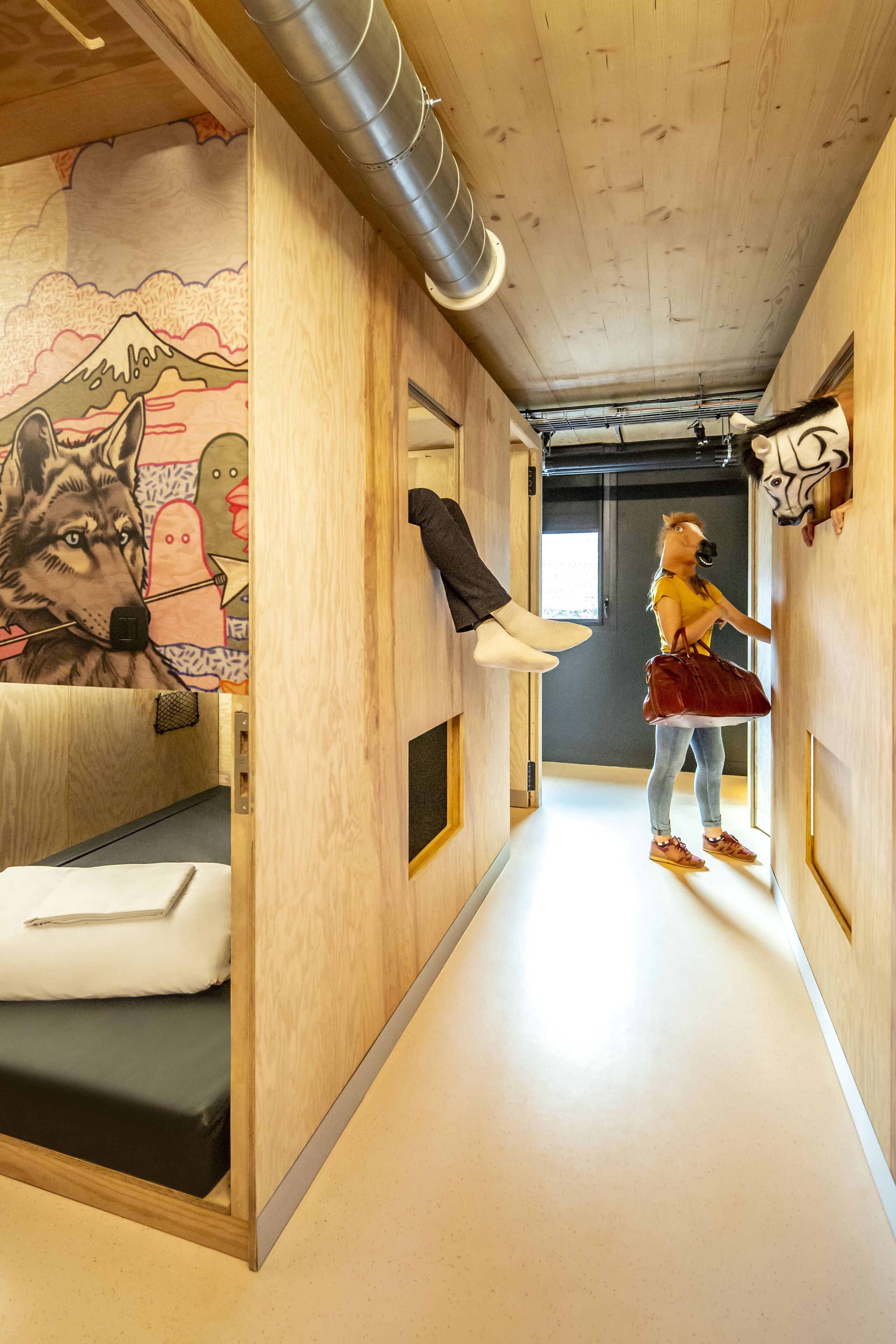 Un pasillo con techo de madera, una cama con colchón blanco, una pared con un mural colorido y una persona de pie cerca de una puerta.