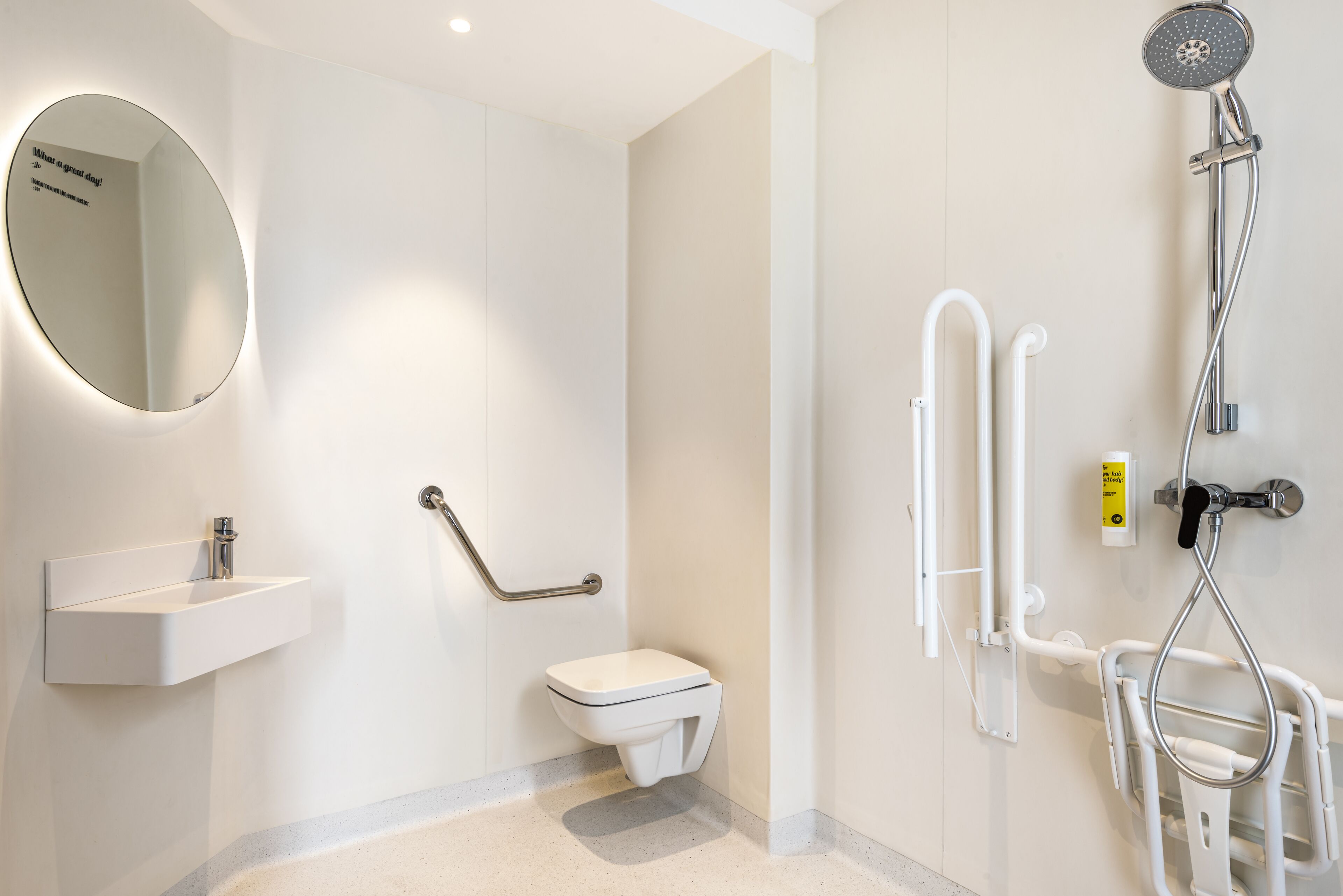 A modern bathroom with a round mirror, a white sink, a toilet, a handheld showerhead, and grab bars.