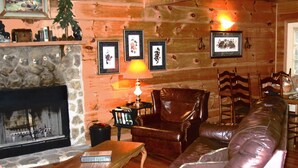 A cozy living room with a stone fireplace, leather sofa, wooden dining table, and framed pictures on the wall. - Clean, Secluded yet Convenient to Town, Well Stocked Cabin in the Woods! Wi-fi! (Townsend)