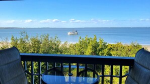 Un balcon avec une table et des chaises, donnant sur une mer calme, avec un bateau visible au loin et une plage en arrière-plan.