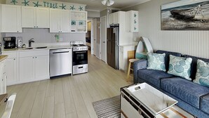 Une cuisine compacte avec des meubles blancs, une cuisinière et un réfrigérateur. On y trouve un canapé bleu avec des coussins à motifs et une petite table.