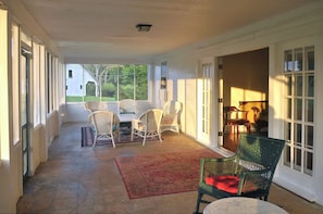 Relax on the covered porch with a set of wicker chairs and a table, surrounded by a large window and a door leading to another room.