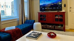 Una sala de estar con un televisor mostrando un paisaje urbano, un aparador rojo con puertas de cristal, un sofá azul y una mesa de centro blanca con un libro y un objeto decorativo.