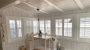 A room with white paneled walls, a wooden ceiling, and a wooden floor. There is a white table with chairs, a hanging light fixture, and a window with blinds.