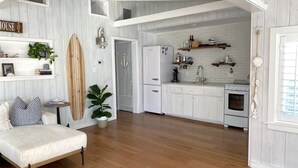 A spacious living area with a sofa, a surfboard, and a kitchen with white cabinets and a refrigerator.