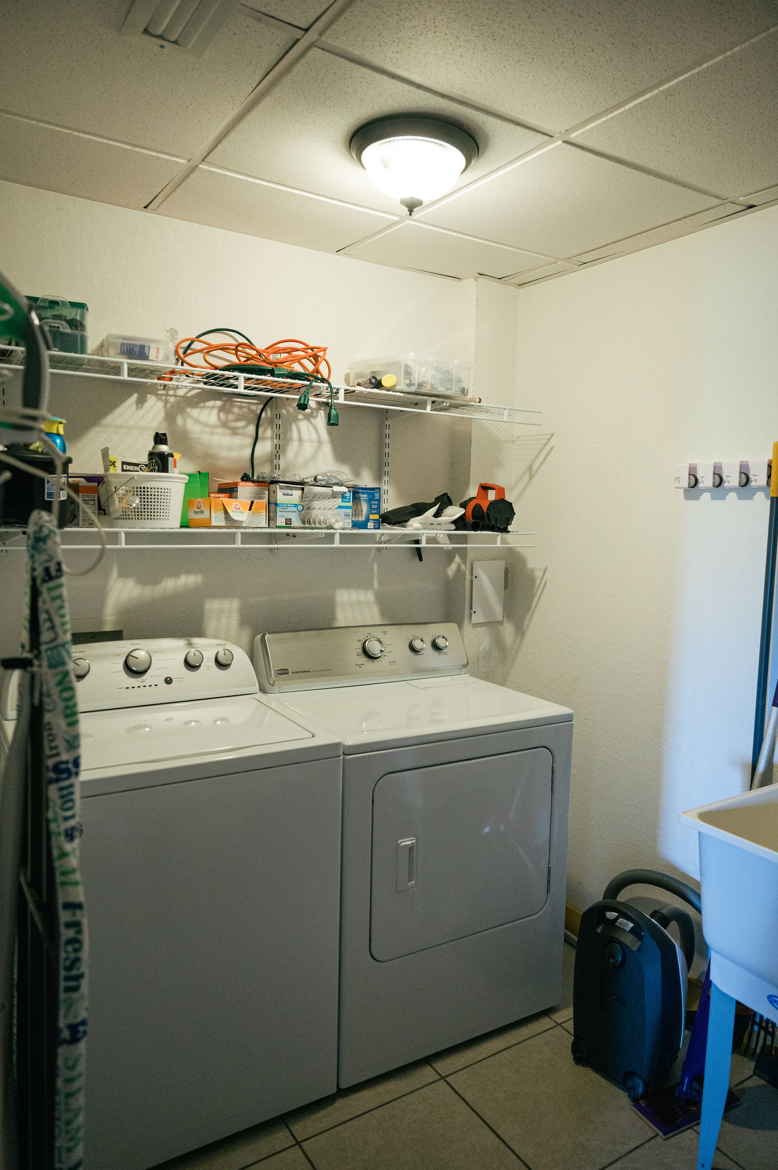 A laundry room with a front-loading washer and dryer, shelves above with various items, and a wall-mounted electrical outlet.