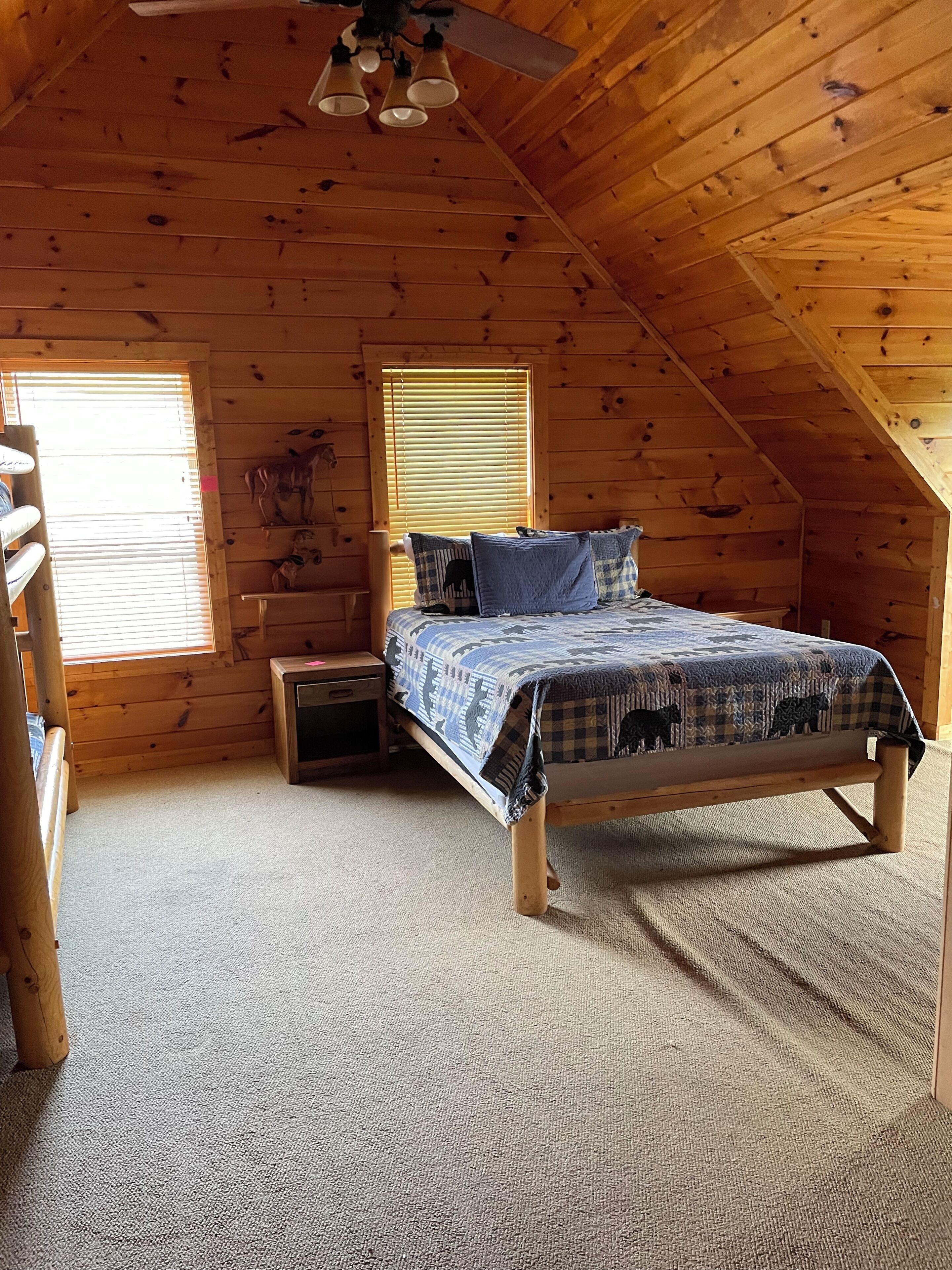 Un dormitorio de cabaña de madera con una cama, una mesita de noche, un aparador, un ventilador de techo y una ventana con persianas.