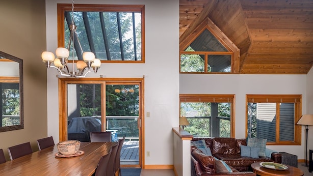 A spacious living area with a wooden dining table, leather sofa, and a large window offering a view of the outdoors. - CEDAR RIDGE 17 | Prime Ski-In, Ski-Out Location | Private Garage & Hot Tub (Whistler)