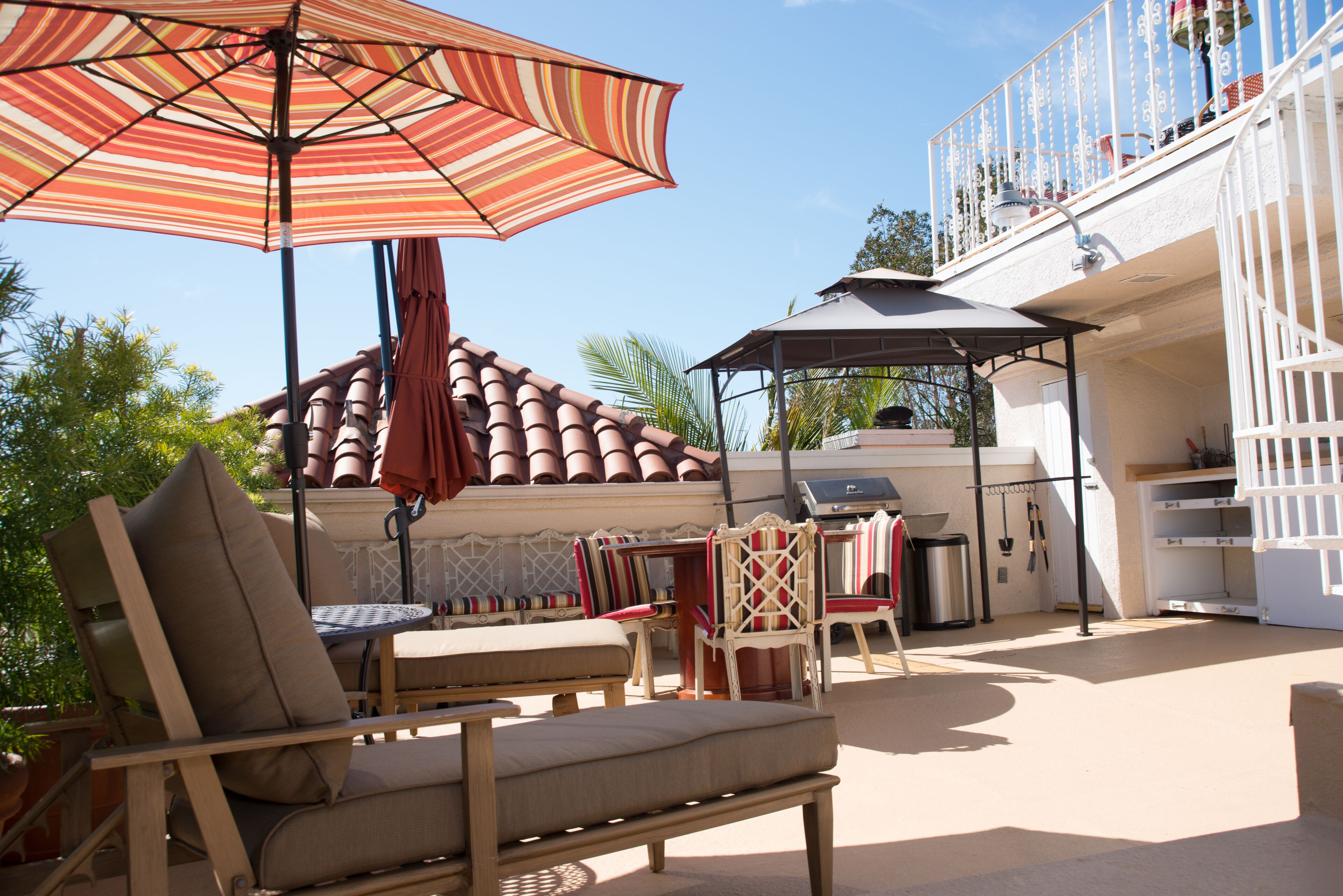 En udendørs terrasse med træmøbler, grill og en stribet parasol.